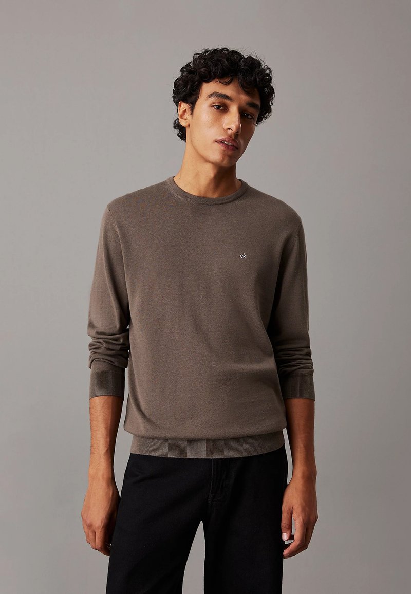 Maglione in maglia marrone con maniche lunghe e scollatura a girocollo. Presenta un piccolo logo bianco sul petto. Abbinato a pantaloni scuri.