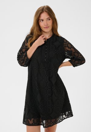 Femme aux cheveux châtain clair portant une robe en dentelle noire à manches trois-quarts avec des boutons sur le devant, debout devant un fond uni.