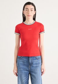 Raudona trumpa rankovėmis marškinėliai su juodai balta dryžuota iškirpte. Ant krūtinės įspaustas Calvin Klein logotipas, derinami su šviesiai mėlynomis džinsais.