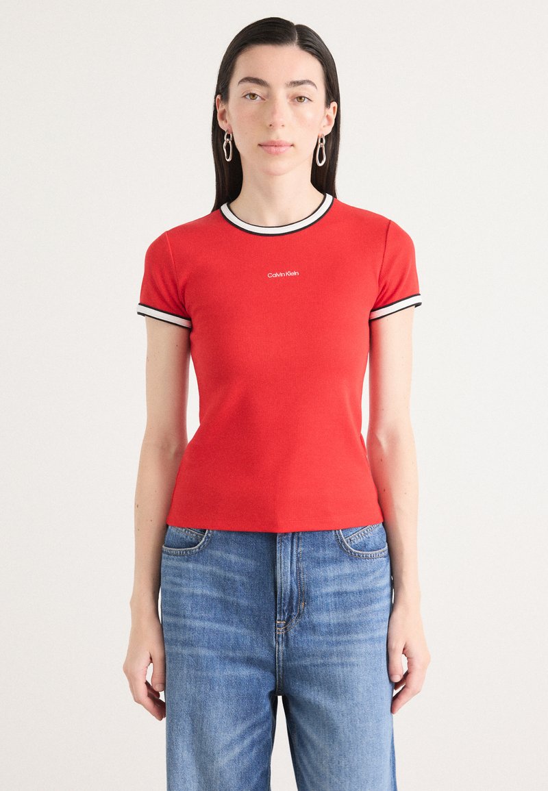Raudona trumpa rankovėmis marškinėliai su juodai balta dryžuota iškirpte. Ant krūtinės įspaustas Calvin Klein logotipas, derinami su šviesiai mėlynomis džinsais.