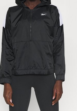 Person trägt schwarze Kapuzenjacke mit Frontreißverschluss und weißen Streifen an den Ärmeln, kombiniert mit schwarzen Leggings.