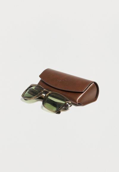 Sončna očala s prozornim okvirjem in zelenimi lečami, ki ležijo ob rjavi usnjeni torbici na belem površju.