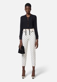 Blusa negra de manga larga con parte frontal con cordones, pantalones blancos con estampado, tacones beige de punta, y un pequeño bolso de mano negro.