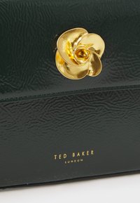 Grön texturerad läderhandväska med ett guldblomformat lås; har präglade detaljer och guldbranding på framsidan.