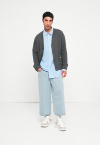 Siv pleteni cardigan s gumbi, svetlo modra majica s črtami, ohlapne svetle jeans hlače in bele superge s bež poudarki.