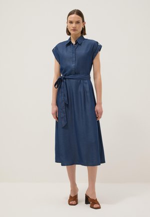 Femme portant une robe midi bleu foncé sans manches avec une ceinture nouée à la taille et des sandales à talons marron, debout devant un fond uni.