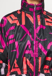Černá zipová bunda s odvážnými abstraktními písmenkovými vzory v růžové a červené barvě a vysokým límcem, kterou nosí osoba.