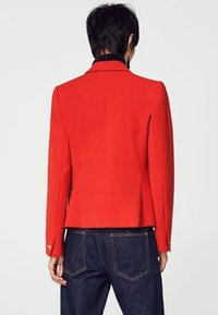 Blazer rosso con un design strutturato, caratterizzato da un ampio colletto e dettagli con bottoni. Realizzato in stoffa testurizzata, ha una vestibilità su misura e uno spacco singolo.