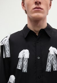 Μαύρο πουκάμισο με λευκές δαντελένιες κορδέλες σε διάφορα μεγέθη, υφασμένη δαντέλα και γιακά με κουμπιά.