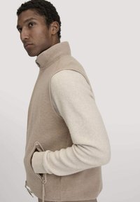 Beige Fleece-Weste mit hohem Kragen und seitlichen Taschen; hat kontrastierende cremefarbene Ärmel und eine glatte Textur. Reißverschlussdetails am Saum.