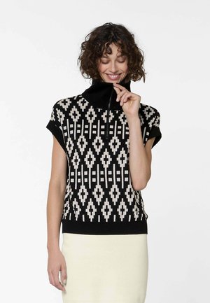 Lächelnde Frau mit lockigem Haar, die einen kurzärmeligen Pullover mit schwarz-weißem, geometrischem Muster, einem Reißverschluss am Kragen und einen leichten Rock trägt.