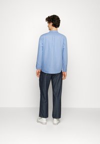 Camisa azul clara de manga longa com pregas nas costas e detalhe de botão, combinada com calças de denim escuras e ténis brancos.