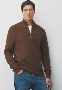 Brauner Strickpullover mit Viertel-Reißverschluss und strukturiertem Muster, über einem weißen Hemd getragen. Das Model posiert mit Händen in den Taschen und trägt eine blaue Jeans.
