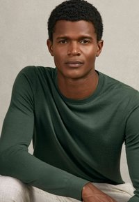 Langärmliges, Rundhalsoberteil in dunklem Grün, glatte Textur, taillierter Schnitt, kombiniert mit hellen, strukturierten Hosen. Neutraler Hintergrund.