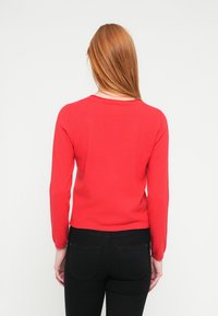 Suéter rojo de manga larga con cuello redondo y puños acanalados, combinado con jeans negros. La textura del suéter parece suave y ajustada.