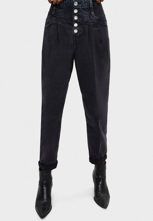 Schwarze, hoch taillierte, schmal zulaufende Jeans mit sichtbaren vorderen Knöpfen, aufgerollten Bündchen und kombiniert mit schwarzen, spitz zulaufenden Stiefeletten.
