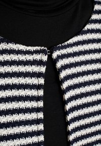 Cardigan a righe con tessuto intrecciato blu navy e bianco, con scollo rotondo e chiusura frontale con gancio in metallo. Strato nero sottostante.