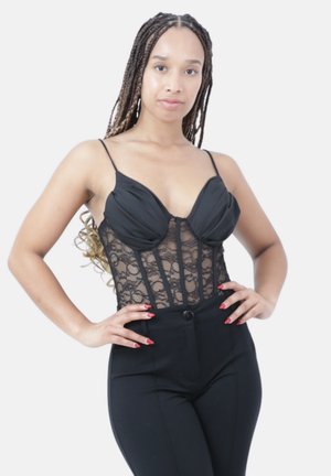 Femme aux longues tresses debout, les mains sur les hanches, vêtue d’un body en dentelle noir et d’un pantalon noir taille haute, faisant face vers l’avant.