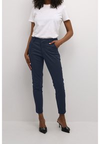 Pantalon bleu marine coupe slim en tissu lisse, avec poches latérales, taille structurée et design fuselé des jambes, associé à des talons noirs.