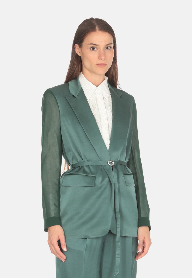 Blazer verde din satin cu mâneci transparente, talie cu curea, două buzunare frontale și guler cu retezare, suprapus peste o bluză albă texturată.