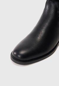 Botín de piel negra hasta el tobillo con punta redondeada, textura suave y diseño minimalista. Cuenta con una suela de goma negra baja.