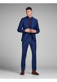Blue tailored suit with notch lapels, slim-fit trousers, and dark button-up shirt, paired with brown leather shoes. Smooth texture and structured design.