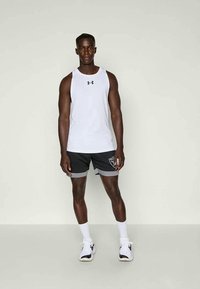 Vyrų sportininkas stovi, vilkėdamas baltą sansleeve Under Armour marškinėlį, juodus ir pilkus šortus su logotipu, baltas kojines ir bėgimo batelius.