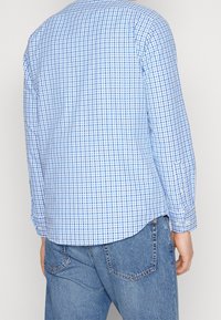 Langarmhemd mit einem blau-weißen Karomuster, abnehmbarem Kragen und abgerundetem Saum. Kombiniert mit hellblauen Jeans.