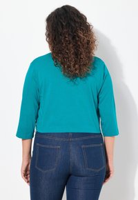 Chemisier teal court à manches trois quarts associé à un jean bleu foncé avec des poches et des détails de couture contrastants.