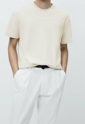 Person wearing a cream short-sleeve t-shirt tucked into high-waisted white pants with hands in pockets, featuring a black belt.