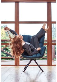 Blaue Stoff-Lounge-Stuhl mit einer gewölbten Sitzfläche, unterstützt von einem schwarzen Metallgestell; eine Person lehnt mit hochgelegten Füßen zurück, umgeben von großen Fenstern.