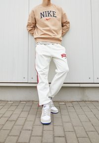 Beige Nike sweatshirt med logotyp, vita sweatpants med röda sidostrikar och Nike-logotyp, samt ljusgrå sneakers med blå accenter.