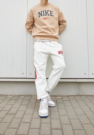 Felpa beige Nike con logo, pantaloni sportivi bianchi con strisce rosse che presentano il logo Nike, e sneakers grigio chiaro con dettagli blu.