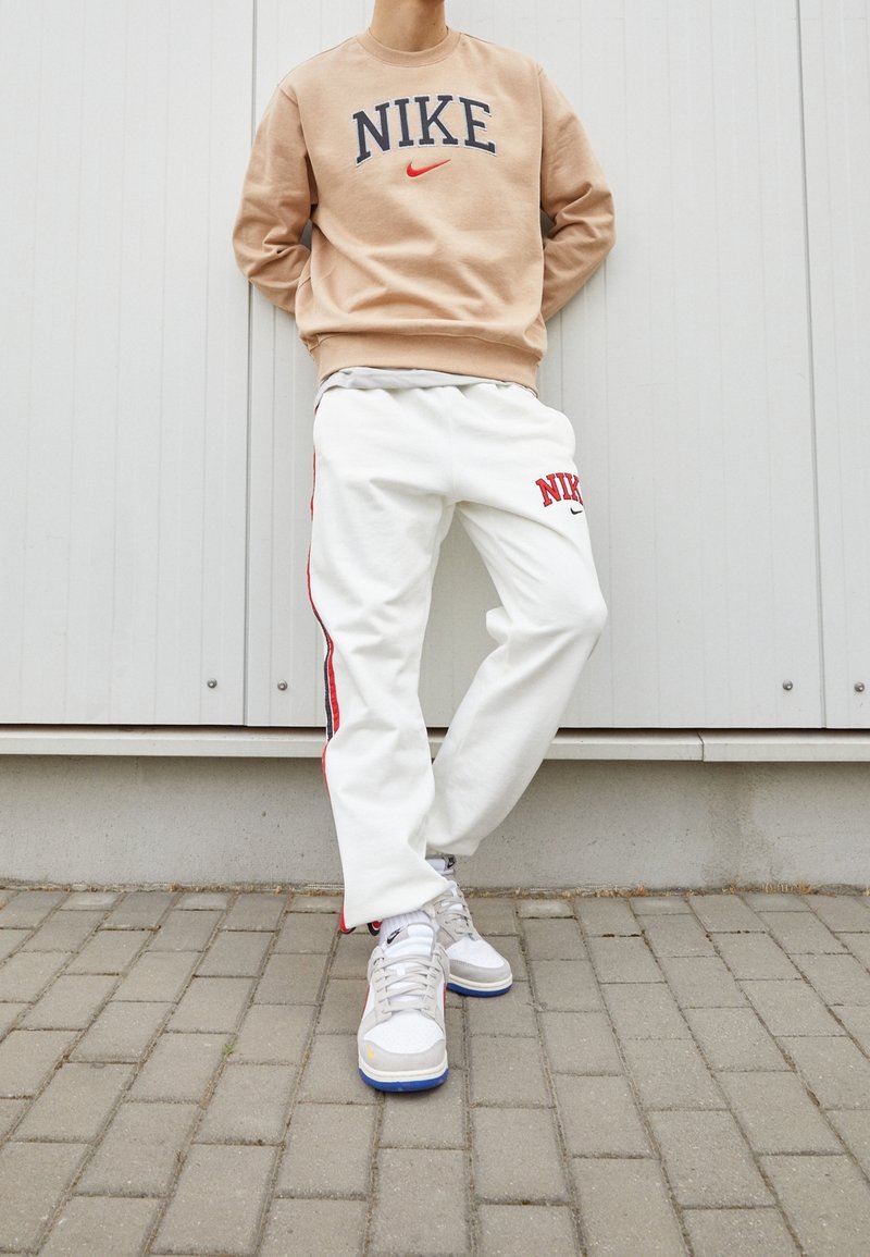 Beige Nike sweatshirt med logotyp, vita sweatpants med röda sidostrikar och Nike-logotyp, samt ljusgrå sneakers med blå accenter.