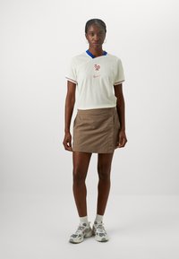 Biela krátka športová košeľa Nike s modrým golierom a červenými akcentmi, s logom kohúta. Hnedá mini sukňa so bočnými gombíkmi. Šedé tenisky.