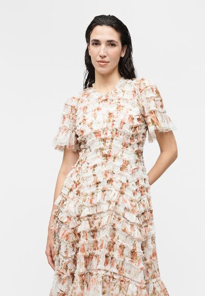 Γυναίκα με σκοτεινά νωπά μαλλιά φοράει ένα λευκό φόρεμα με λουλούδια, με στρωτές βολάν και κοντά φουσκωτά μανίκια, στέκεται μπροστά σε απλό φωτεινό φόντο.