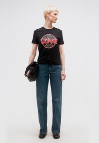 Άτομο με κοντά ξανθά μαλλιά που φοράει μαύρο t-shirt με γράφημα «LOVE», μπλε τζιν, κρατώντας μαύρη τσάντα, στέκεται σε απλό φόντο.