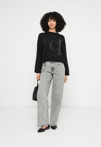 Μαύρο κοντό πουλόβερ με μεγάλο γκρι λογότυπο, σε συνδυασμό με ανοιχτό γκρι τζιν με φαρδύ πόδι. Μαύρη τσάντα χειρός και μυτερά επίπεδα παπούτσια ολοκληρώνουν το ντύσιμο.