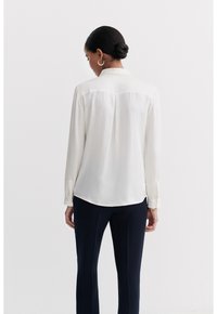Silk blouse in off-white, featuring long sleeves, a subtle back seam, and a curved hem. Paired with navy trousers.