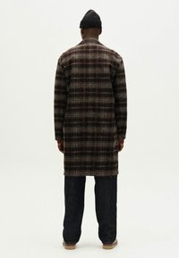 Lang ternet frakke i en mørk farvepalet med brune og grå nuancer. Har en lige skæring og sidelommer. Sættes sammen med mørke jeans og en hue.