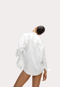 Camisa de lino blanca con un corte relajado, mangas largas y un dobladillo curvado. Detalle de yugo en la parte posterior visible, tejido ligero con un acabado texturizado.