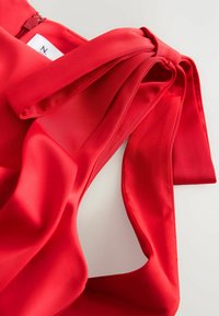 Detail van een rode zijden blouse met een gestrikte strik op de schouder, een gladde textuur en een verborgen rits aan de zijkant.