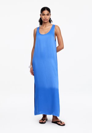 Femme portant une robe maxi bleue sans manches avec des fentes latérales et des tongs noires, debout avec une main derrière le dos.