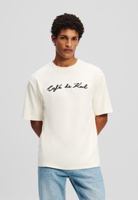 Cream-colored t-shirt made of smooth fabric, featuring short sleeves and a crew neckline, decorated with black cursive "Café de Kal" text.