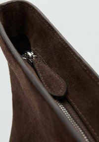 Wnętrze brązowej torebki z zamszu z widocznym srebrnym zamkiem błyskawicznym, gładką podszewką i małym skórzanym uchwytem. Widoczne są szczegóły miękkiej tekstury.