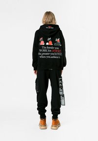 Persona de pie de espaldas vistiendo una sudadera negra con texto y gráficos rojos y blancos, pantalones cargo negros con correas y botas beige.