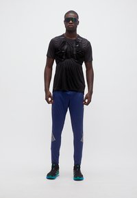 Άνδρας που στέκεται φορώντας μαύρο αθλητικό μπλουζάκι με γιλέκο ενυδάτωσης, μπλε φόρμες, μαύρα παπούτσια τρεξίματος και ανακλαστικά αθλητικά γυαλιά ηλίου.