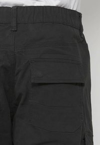 Zwarte cargobroek met elastische tailleband, meerdere zakken en sterke stiksels. De stof lijkt stevig en heeft een gladde textuur.