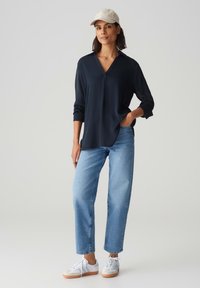 Blusa navy a scollo a V con maniche lunghe, abbinata a jeans dritti azzurri e sneakers bianche con suole beige, e un cappellino beige.