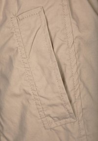 Beige tyg med en rektangulär lapplåda, med prydlig sömnad och en slät yta. Fickan är placerad på sidan.